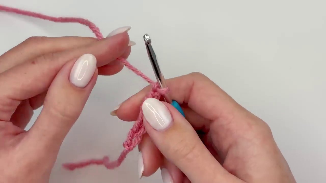 The hook demonstrates a triple crochet stitch being worked into the center of the ring, with four loops visible on the hook.