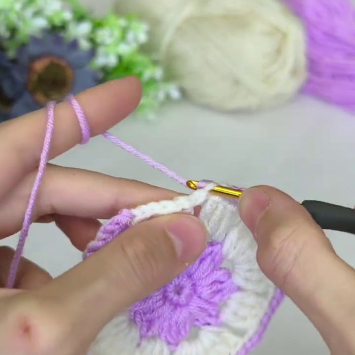 The crochet hook and light purple yarn sit ready on a neutral surface, with a ball of white yarn visible in the background.