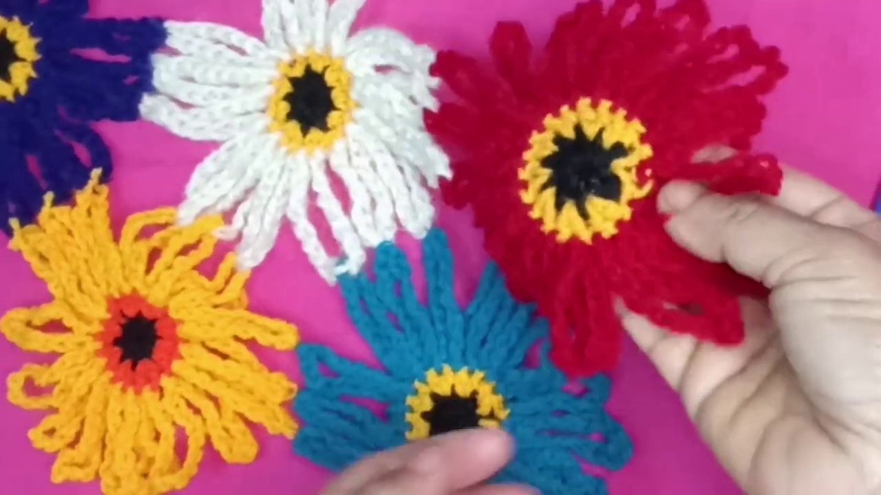A close-up of the white crochet hook working through black yarn, with colorful flowers scattered in the background.