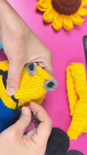 The maker’s hands sewing the eye and mouth pieces onto the yellow body, with the pink background visible.