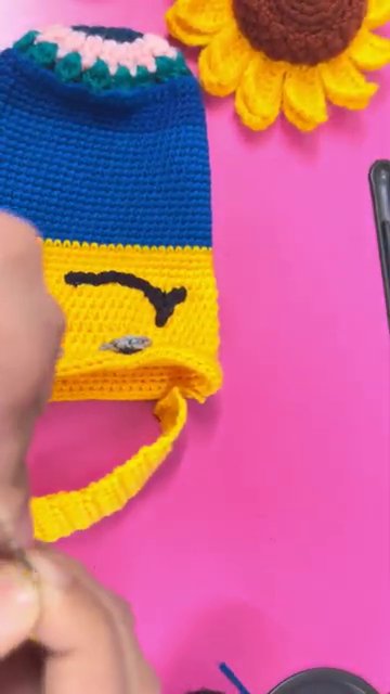 The bottle cover sitting next to a crocheted sunflower on the pink work surface, showing the scale of the finished piece.