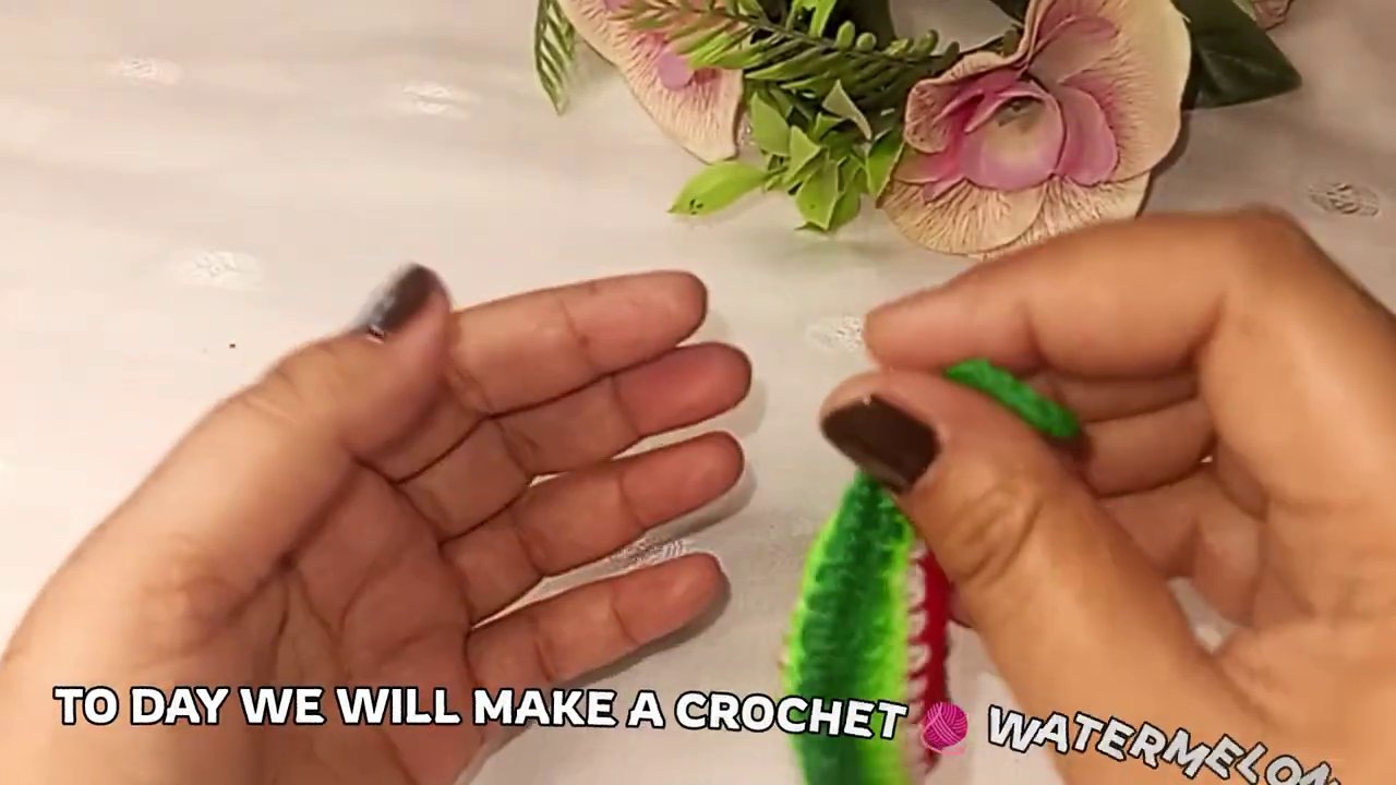 Red yarn and crochet hook on a white tablecloth, with green yarn and a small bouquet of flowers visible in the background.