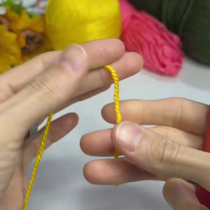 Yarn balls in yellow, pink, and green sit on a white table beside a red-handled crochet hook.