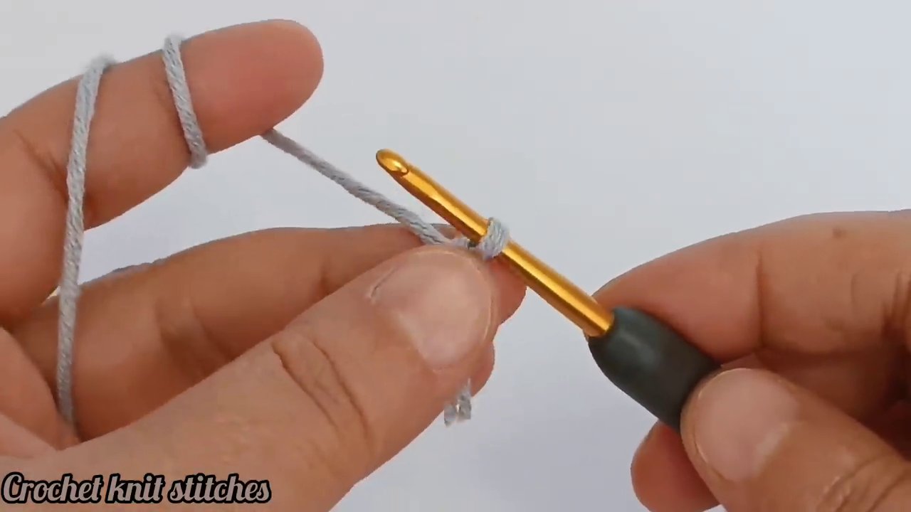 Hands stretch a length of yarn between thumbs and index fingers, creating tension. A loop of yarn sits in the background as the hook prepares to enter the work.