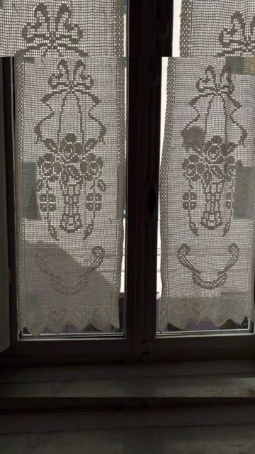 Measuring a window with a tape measure, a partially finished crochet curtain draped over the rod for reference.