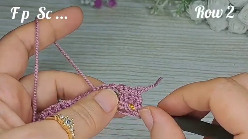 The full workspace: a light wooden surface, pink yarn, gold hook, and a small bunch of white flowers in the upper right corner.