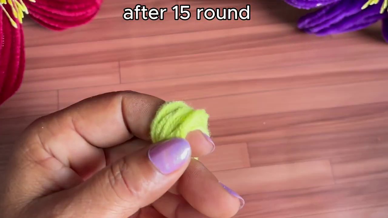 Hands with light purple nail polish hold yellow yarn and a white crochet hook against a wooden work surface.