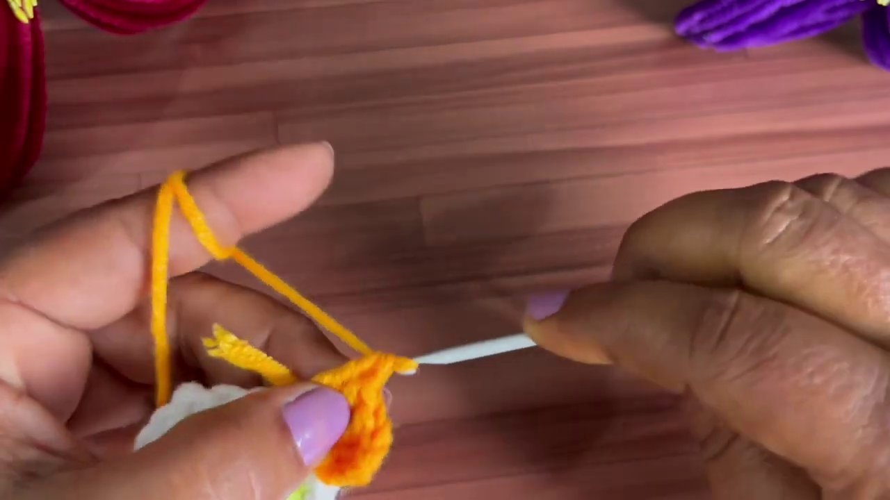A close-up shows orange yarn being pulled through a stitch to form the first petal curve.